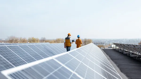 Rows of solar panels on a large rooftop with two workers in hard hats walking between rows.