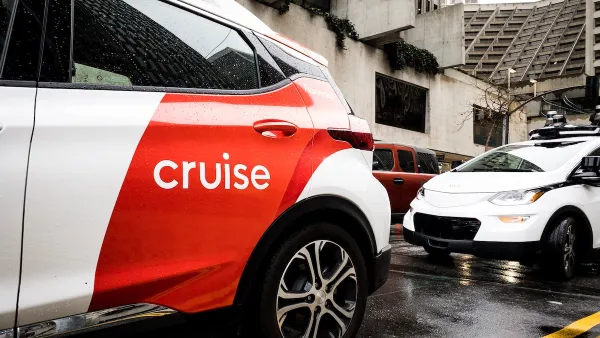White and red Cruise autonomous car on San Francisco street with concrete Brutalist-style building in background on grey, rainy day.