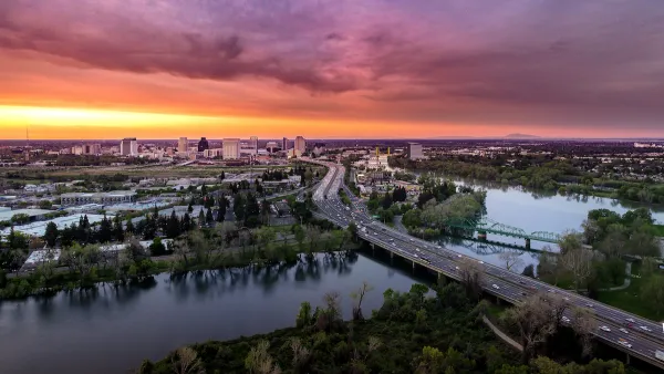 Aerial view of freeway crossing river in Sacramento, California with colorful sunset sky in background.