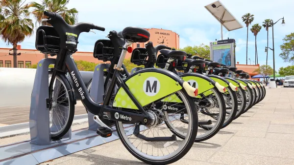 Row of black and green Los Angeles Metro bike share bikes at station with red building with clock and'Los Angeles' lettering in background.