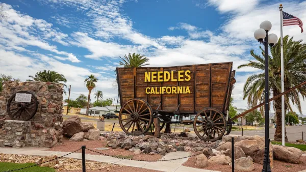 Sign for Needles, California installed on wooden wagon with residential neighborhood visible in background.
