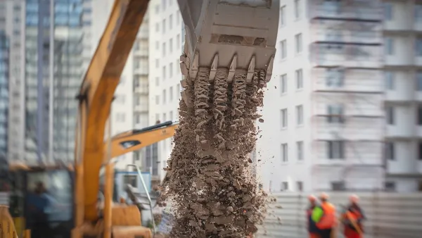 Excavator digging in an urban street with two workers standing next to it.