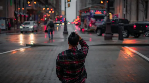 View from behind of person standing on city sidewalk wearing red and black plaid shirt looking across street at dusk.