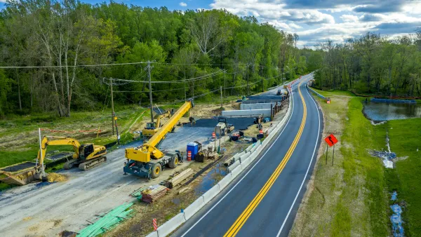 Aerial view of road construction with yellow heavy equipment parked next to two-lane asphalt road against green forest and blue sky