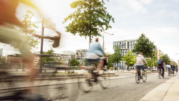 Line of five cyclists bike away from camera in wide protected bike lane on the right side of a street, with low-rise residential buildings in the background.