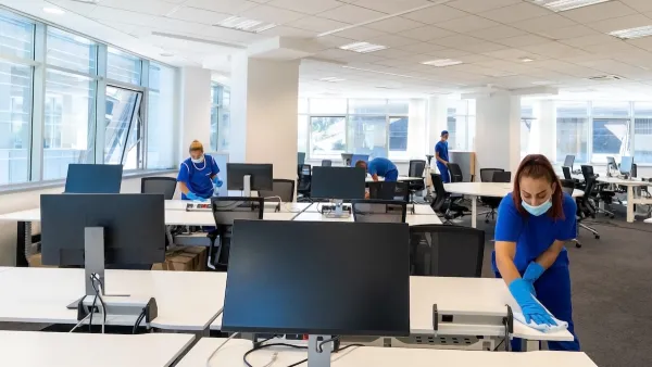 Professional cleaners wearing medical masks and gloves cleaning desks in large empty office.