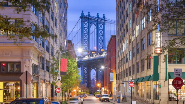 View of Brooklyn Bridge at night from Brooklyn alley with multi-story buildings and parked cars.