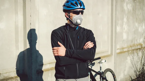 Male cyclists wearing helmet, sunglasses, and N95 mask stands with arms crossed in front of his bike
