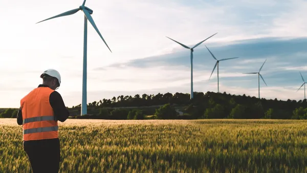 Technician in orange vest and white hard hat standing in field with row of wind turbines.