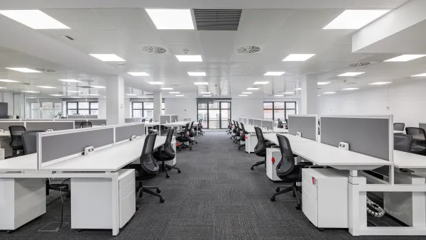 Large empty open-plan office with rows of white desks and black chairs under bright fluorescent lighting