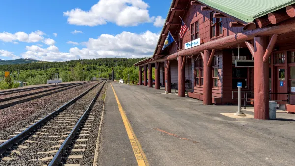Rail tracks on the left, rustic log-built train station painted reddish brown with a green metal roof and concrete platform on the right, evergreen forest and bright blue sky with fluffy white clouds in the background.