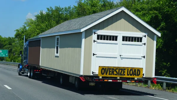 Tan modular building with white swinging barn/garage door being transported on freeway with yellow "Oversize Load" banner across back.