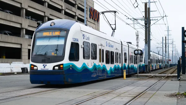 Link Light Rail train travels on ground level tracks paralell to a street with electrical wires overhead.