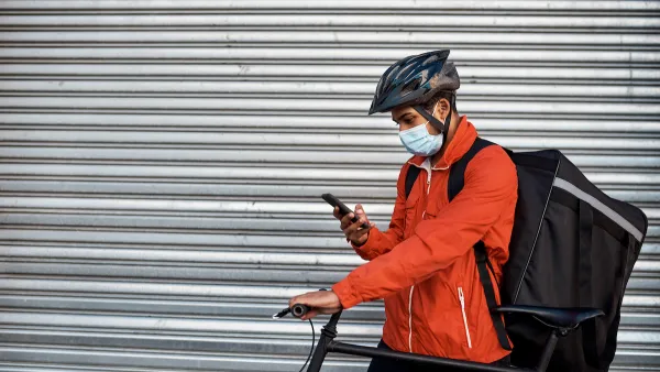 Man in orange jacket wearing surgical mask and bike helmet looks at phone while wearing delivery backpack and holding bicycle