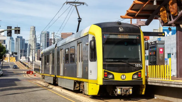 Yellow and black light rail train at street-level station with downtown Los Angeles skyline in background.
