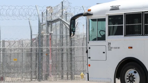 White prison transport bus passing in front of heavily fortified prison fence with barbed wire.