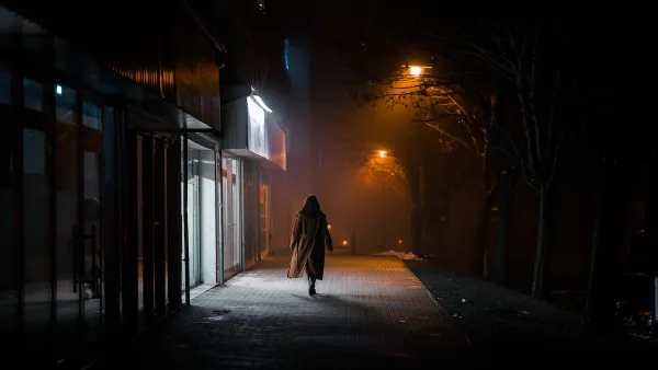 Silhouette of woman in long coat walking on darkened city street alone at night, passing brightly lit sotrefront.