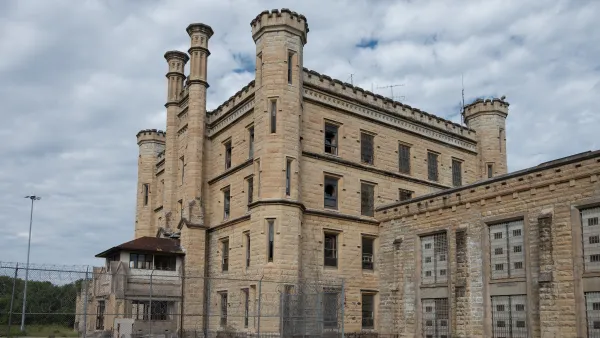 Deralict stone prison building with turrets and a chain link fence in foreground, under a gray, cloudy sky.
