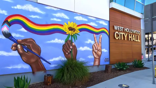 Front entrance of West Hollywood City Hall with name on side of building and mural of hands painting a rainbow across a blue sky.