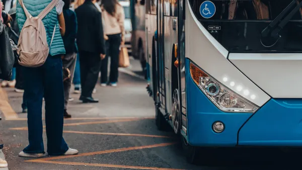 Close-up of front side of blue and white bus with accessibility sticker and people wearing backpacks waiting outside of bus.