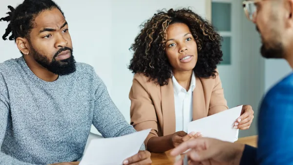 A Black couple holding contracts sits across the desk from a white mortgage lender.