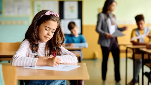 Young girl sitting at school desk taking test with blurred teacher and other students in background.