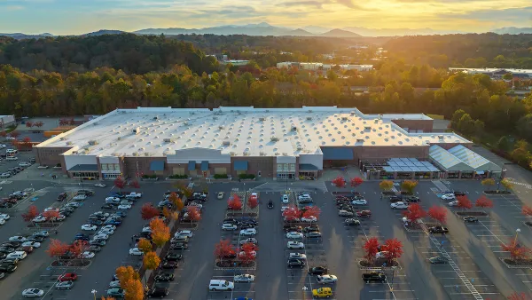 Aerial view of large parking lot in front of large grocery or big-box store with small fall-colored trees among parked cars and sunset sky in background.