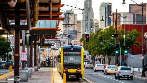 Yellow and black Metro light rail train pulling up to outdoor platform with Los Angeles skyline in background.