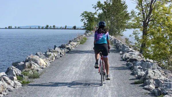 View from behind of woman wearing helmet on bicycle riding on concrete causeway with water on either side.