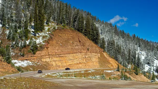 A road winds along the edge of a mountain, with an inclined tan rockface above it and a snowy treeline near the top of a photo.