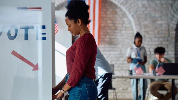 A side view of a young Black woman in a red sweater and jeans standing at a ballot box in a gymnasium with white walls and an American flag in the background.