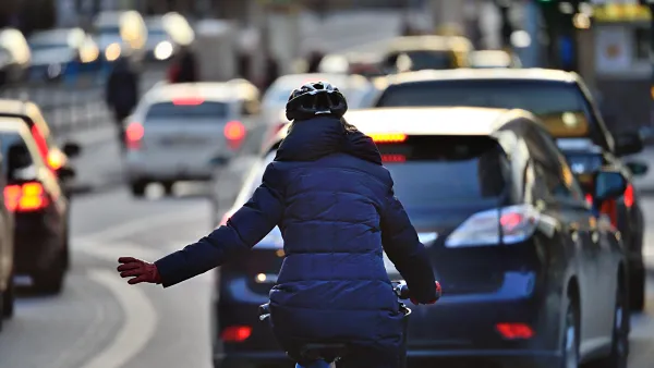 Rear view of woman in blue coat riding bike on busy road signals with left hand to merge into lane of traffic.
