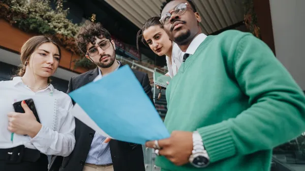 Group of four young professional people standing in front of urban building looking at documents.