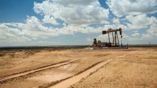 Distant view of brown oil pump jack in a flat desert with cloudy blue sky.