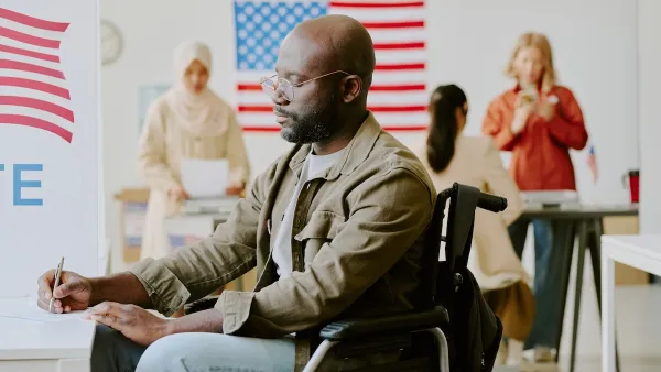 African American man in a wheelchair filling out paper ballot at a polling station with American flags in background.