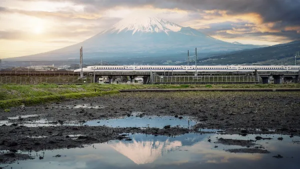 White Shinkansen high-speed rail train passing through green fields with water and Mount Fuji in background in Japan.