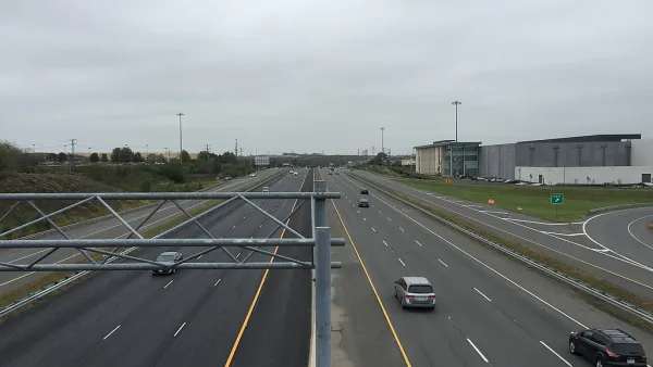 A wide highway with a few cars on a gloomy day. Low-rise commercial buildings and more car infrastructure is visible to the side of the road.