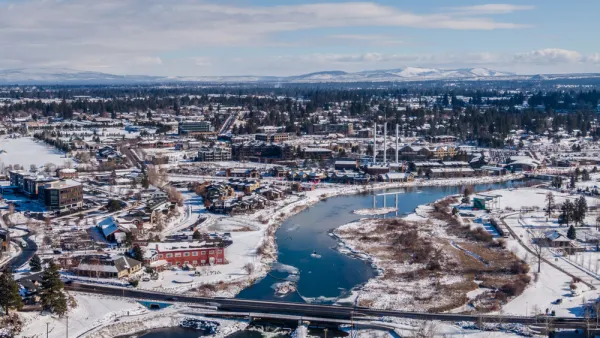 An aerial image of Bend, Oregon, covered in snow in the winter. The Deschutes River cuts through the middle of town, below an old mill.
