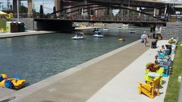 A canal under a bridge in Buffalo is lined by chairs and tables, people walking about, looking at peddle boats on the water.