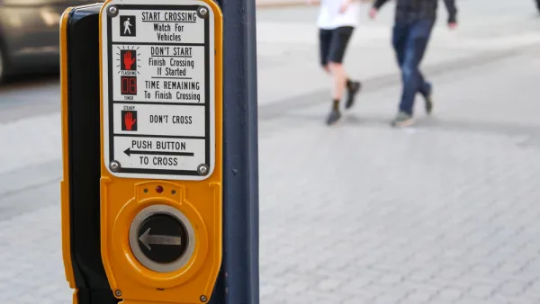 A crosswalk activation button is shown in the foreground with two pedestrians show blurred while crossing the street in the background.