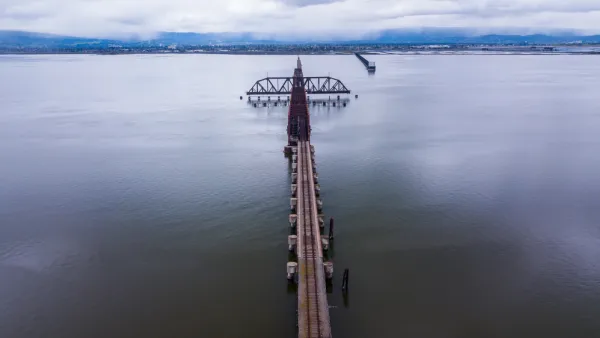 A long rail bridge, rusted and old, crosses a long stretch of water with hills and clouds on the other side.