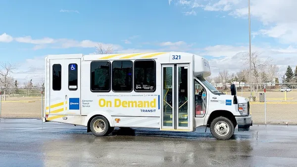 White minibus with yellow "On-Demand" logo parked in an asphalt lot with grass field and dry fall trees in background in Edmonton, Canada.