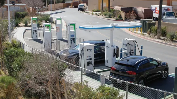 A pair of electric cars plugged into charge at facilities in the corner of a shopping center parking lot.