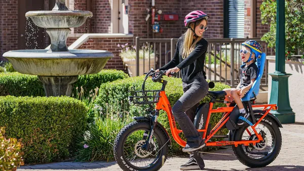 Woman in purple helmet on orange e-bike with baby in seat on back of bike.