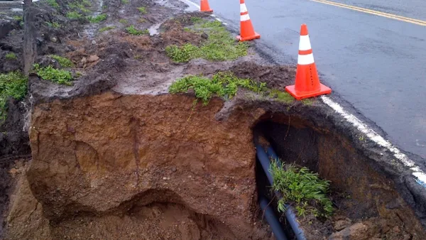 Eroded cliffside and exposed utilities along asphalt Highway 1 in California with orange traffic cones lining side of road.
