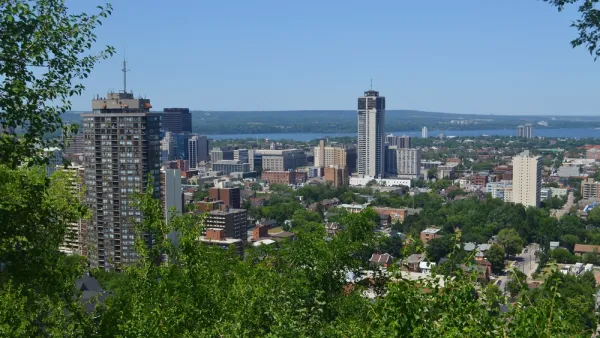 The downtown high rises of Hamilton, Ontario are surrounded by greenery and the blue of Lake Ontario on a sunny day.