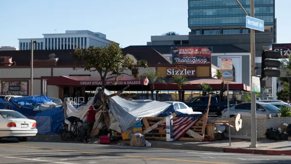 A homeless encampment fills a sidewalk in front of a Sizzler restaurant advertising Thanksgiving dinner.