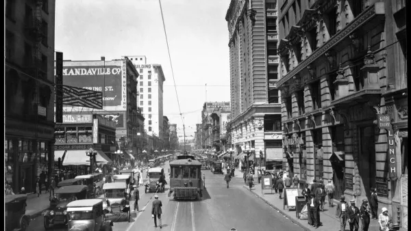 Black-and-white 1924 photo of a street in Los Angeles with a street car, cars driving in the other lane over street car tracks and pedestrians walking on sidewalk and middle of street.