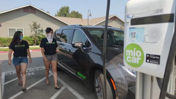 Black SUV charging at Miocar electric car sharing station with two women walking towards it.