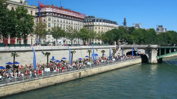 A large crowd gathers on the banks of the River Seine on a sunny day in Paris.